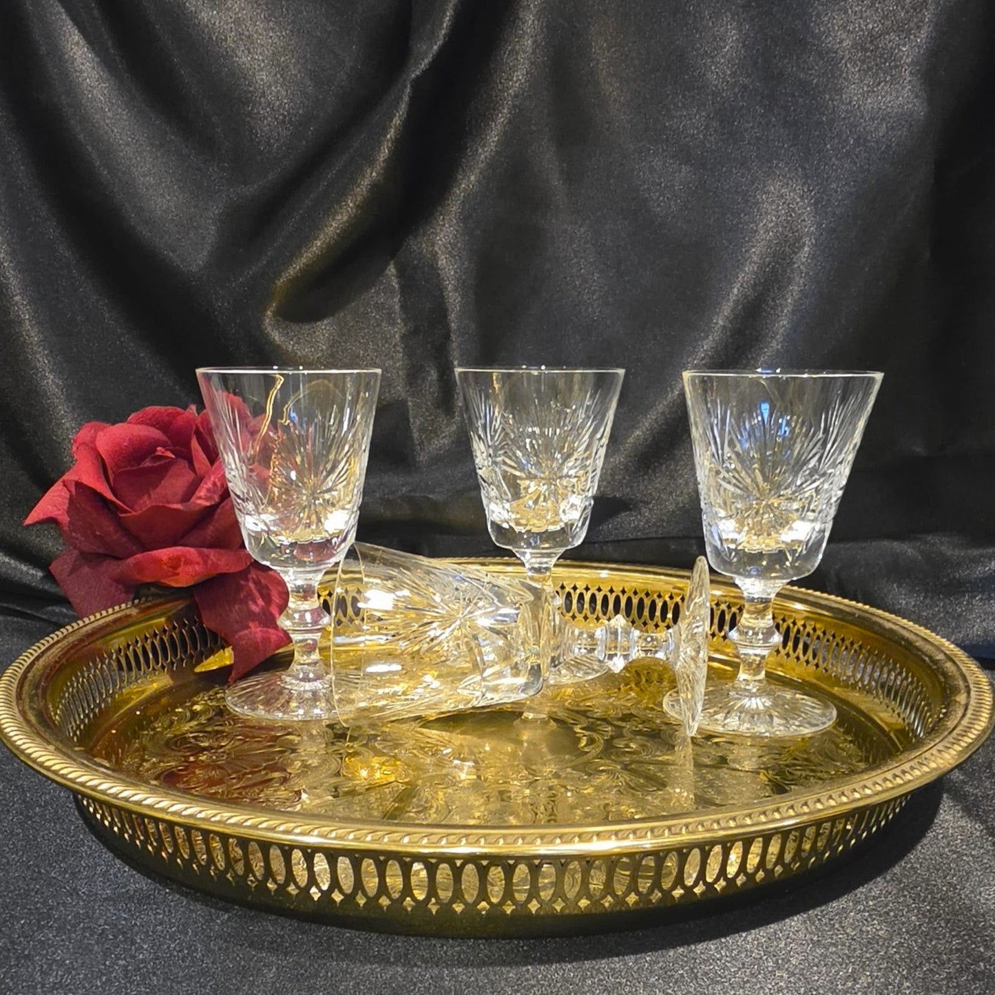 Three crystal glasses on a gold tray with a red rose against a dark fabric background